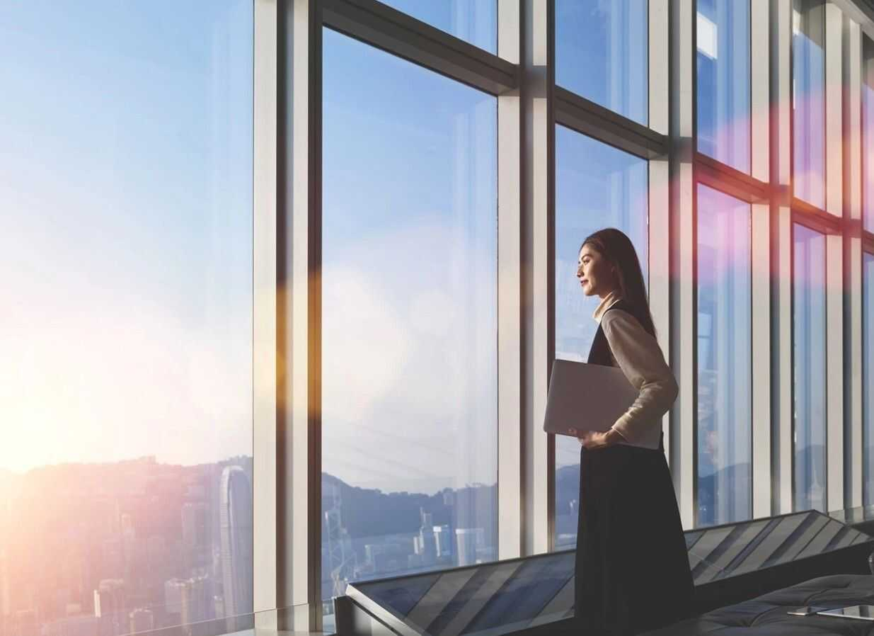 Žena stojaca bokom držiaca laptop pozerajúca cez panoramatické okná