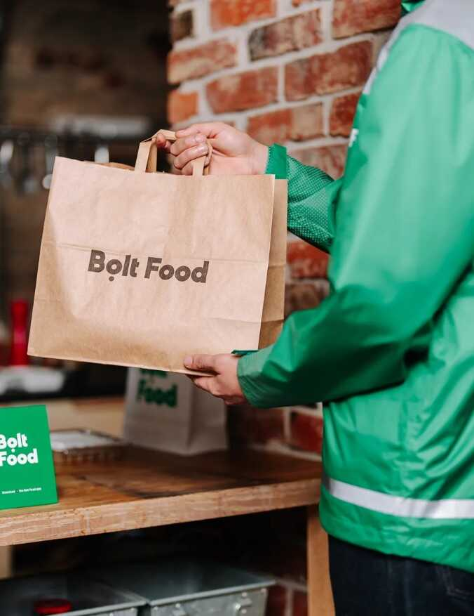 Bolt food kuriér v zelenej bunde odovzdávajúci papierovú tašku
